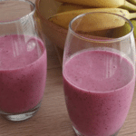 Two tall glasses filled with a finished purple acai smoothie, with a bowl of fresh bananas in the background.