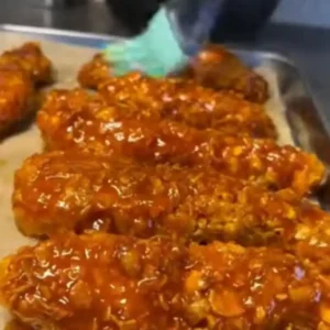 A hand using a pastry brush to apply a shiny, reddish-brown honey-hot glaze to freshly cooked crispy chicken crispers on a baking sheet.