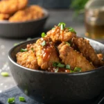 A close-up, professional photograph of golden-brown crispy chicken crispers piled in a dark ceramic bowl, glistening with a shiny honey-hot sauce glaze and garnished with fresh green chives.